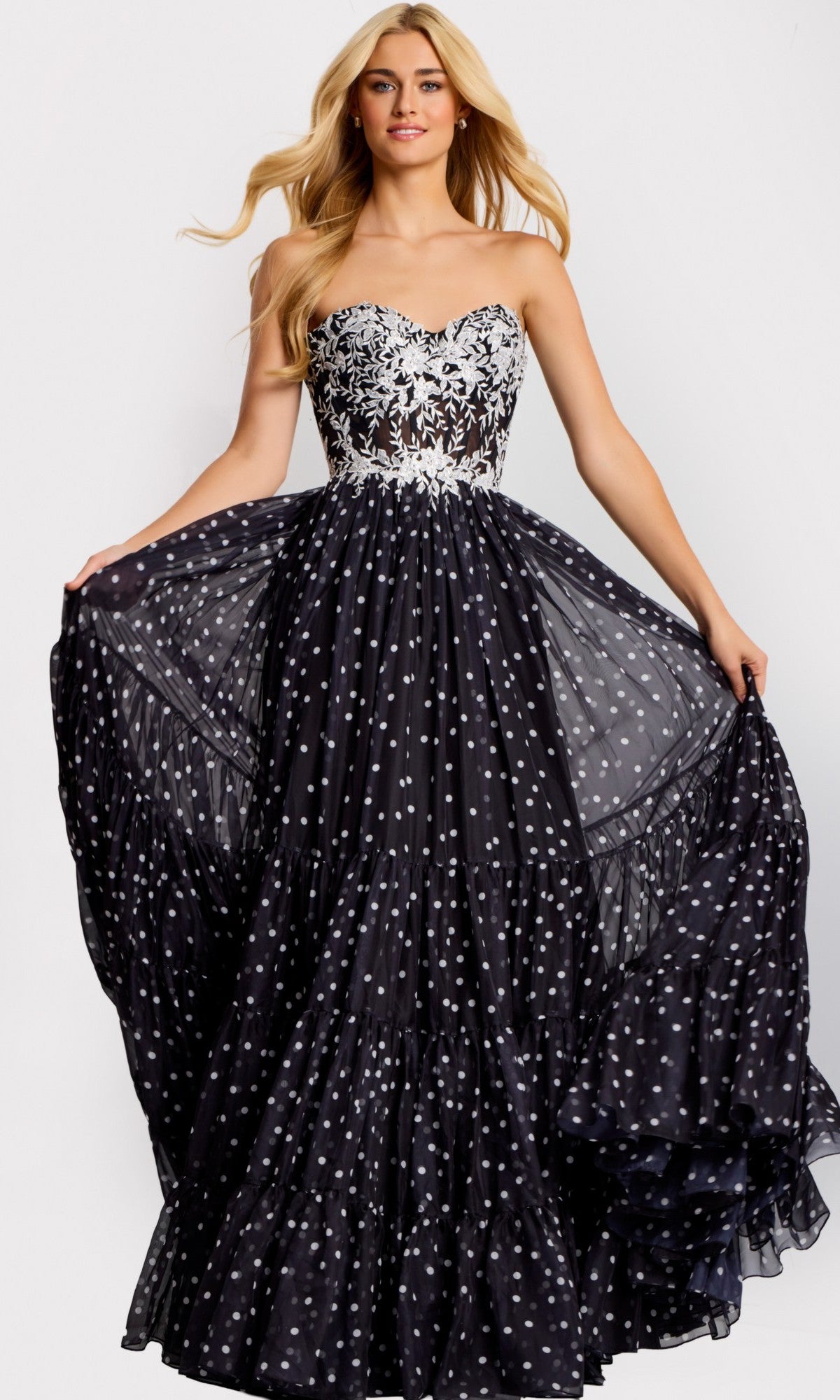 A woman wearing the black Jovani 49086 long prom dress, featuring a strapless floral lace corset bodice and a full, flowing A-line skirt with scattered white polka dots.